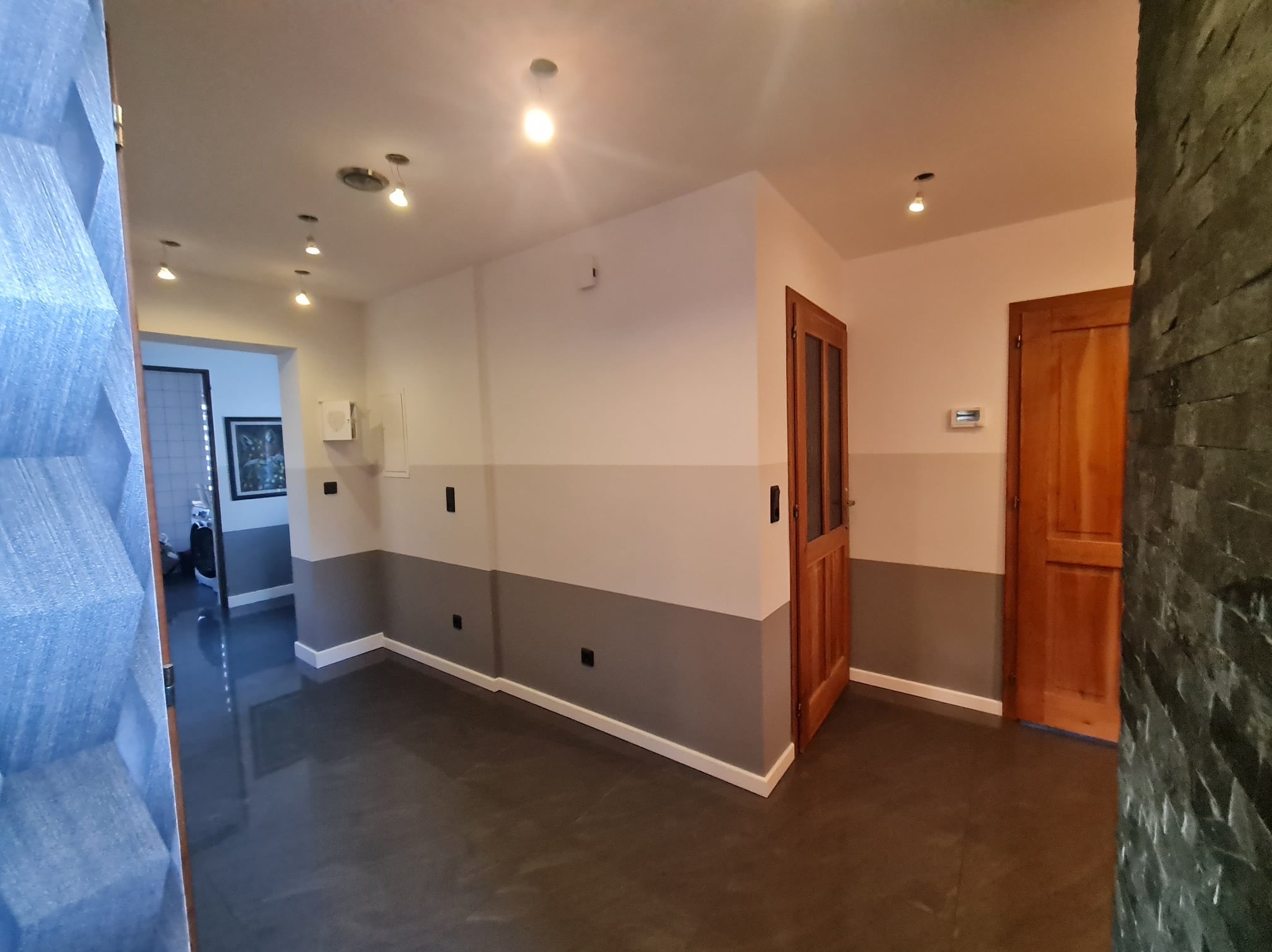 Dwukolorowe ściany w korytarzu z ciemną, błyszczącą podłogą i drewnianymi drzwiami, widok zza dekoracyjnej ściany z kamienia.