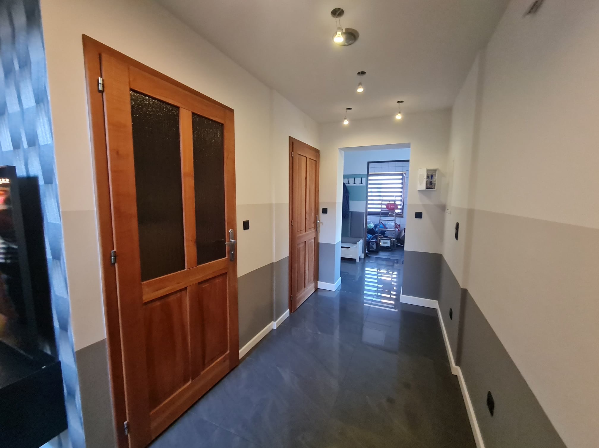 Wykończony korytarz z dwiema drewnianymi drzwiami z przeszkleniami, szarą lamperią na ścianach i ciemną, błyszczącą podłogą z płytek, widok na dalsze pomieszczenie z roletami w oknie.