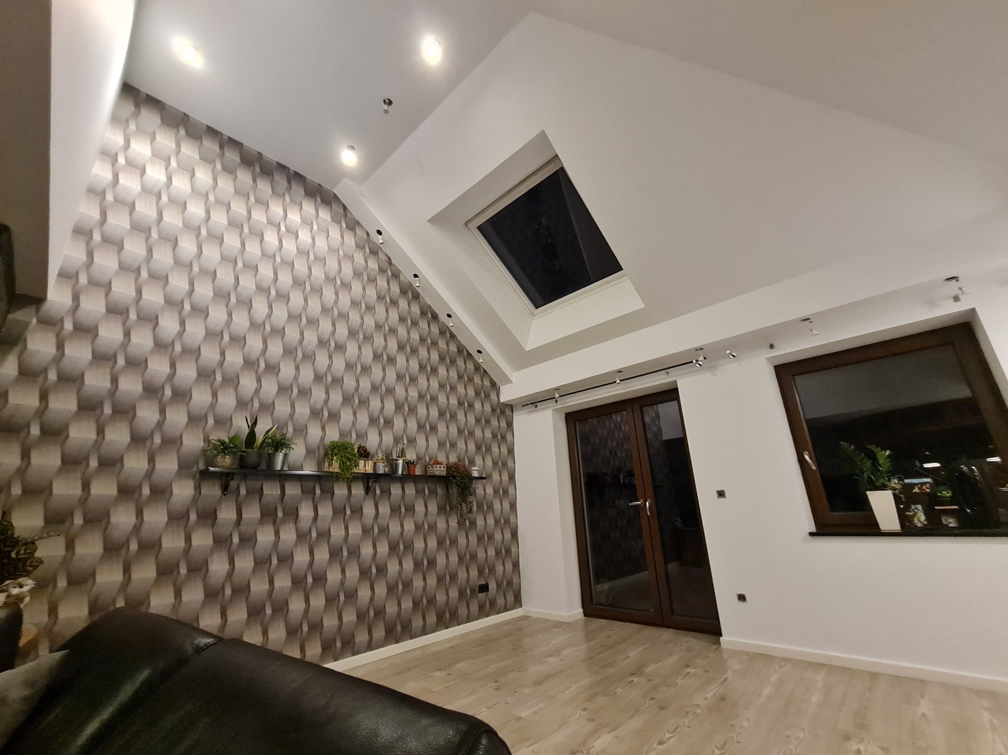 Pokój na poddaszu z oknem dachowym, teksturowaną ścianą, półką z roślinami, drewnianą podłogą i ciemnymi drzwiami balkonowymi.