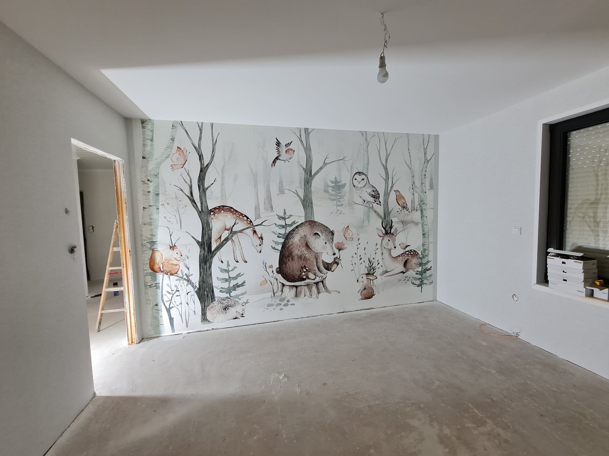 Pokój po remoncie z fototapetą przedstawiającą leśne zwierzęta na jednej ze ścian, surowy betonowy podkład pod wylewkę, widoczna drabina w przejściu do innego pomieszczenia.