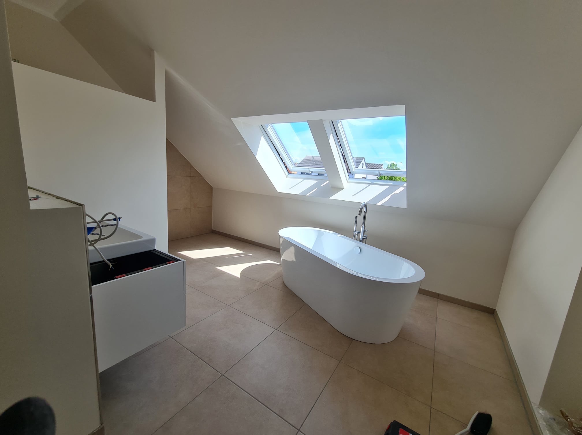 Nowoczesna łazienka na poddaszu z wolnostojącą białą wanną, jasnymi płytkami podłogowymi i trzema oknami dachowymi wpuszczającymi naturalne światło.