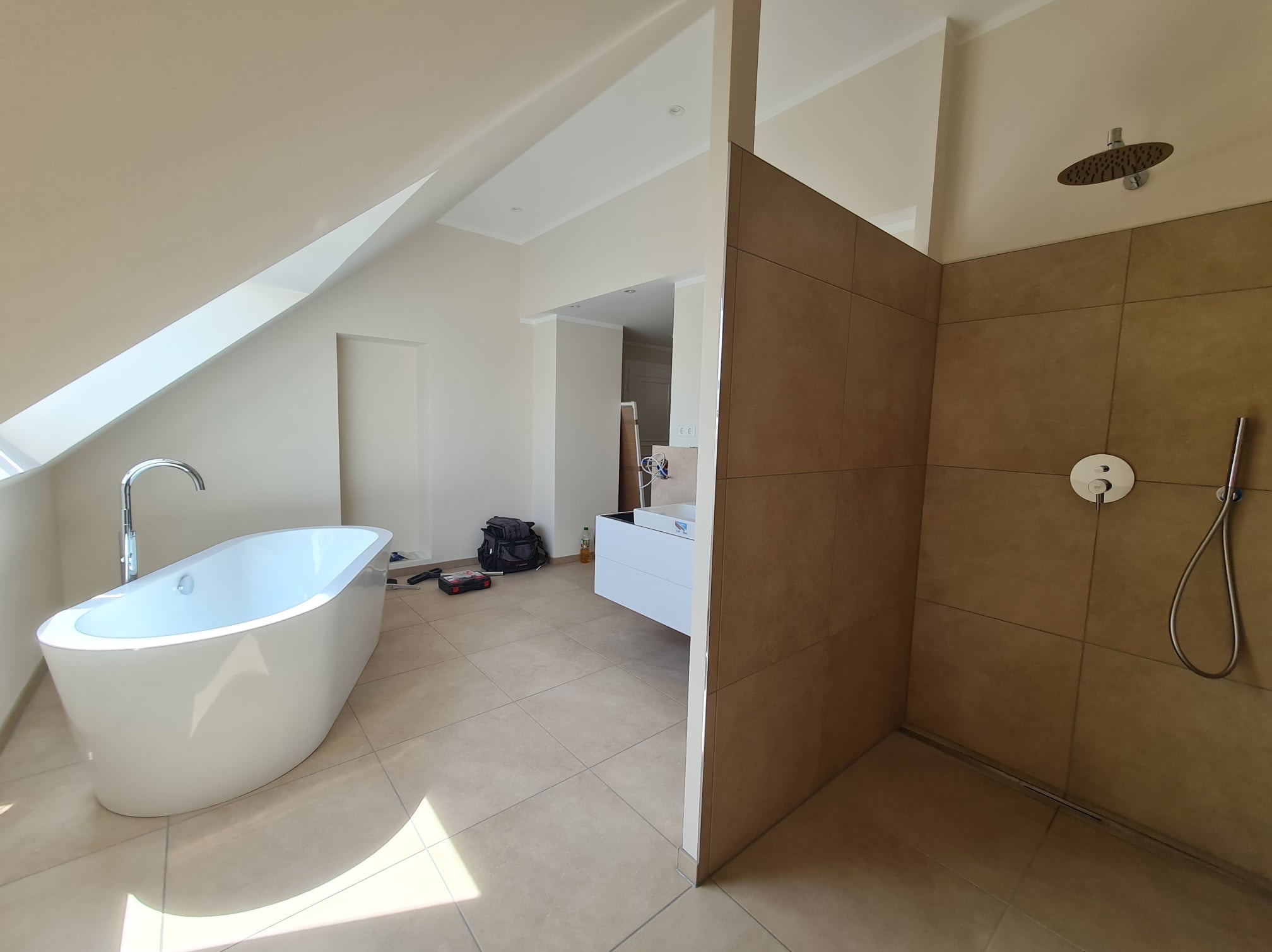 Nowoczesna łazienka w trakcie remontu: wolnostojąca wanna z chromowaną baterią, kabina prysznicowa wyłożona brązowymi płytkami, biała szafka z umywalką, narzędzia i torba monterska na podłodze...