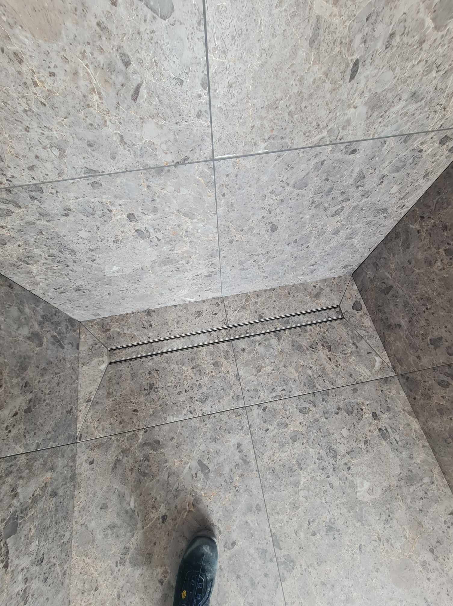 Szare płytki imitujące marmur na podłodze i ścianie, widoczny odpływ liniowy i fragment buta w dolnym rogu.