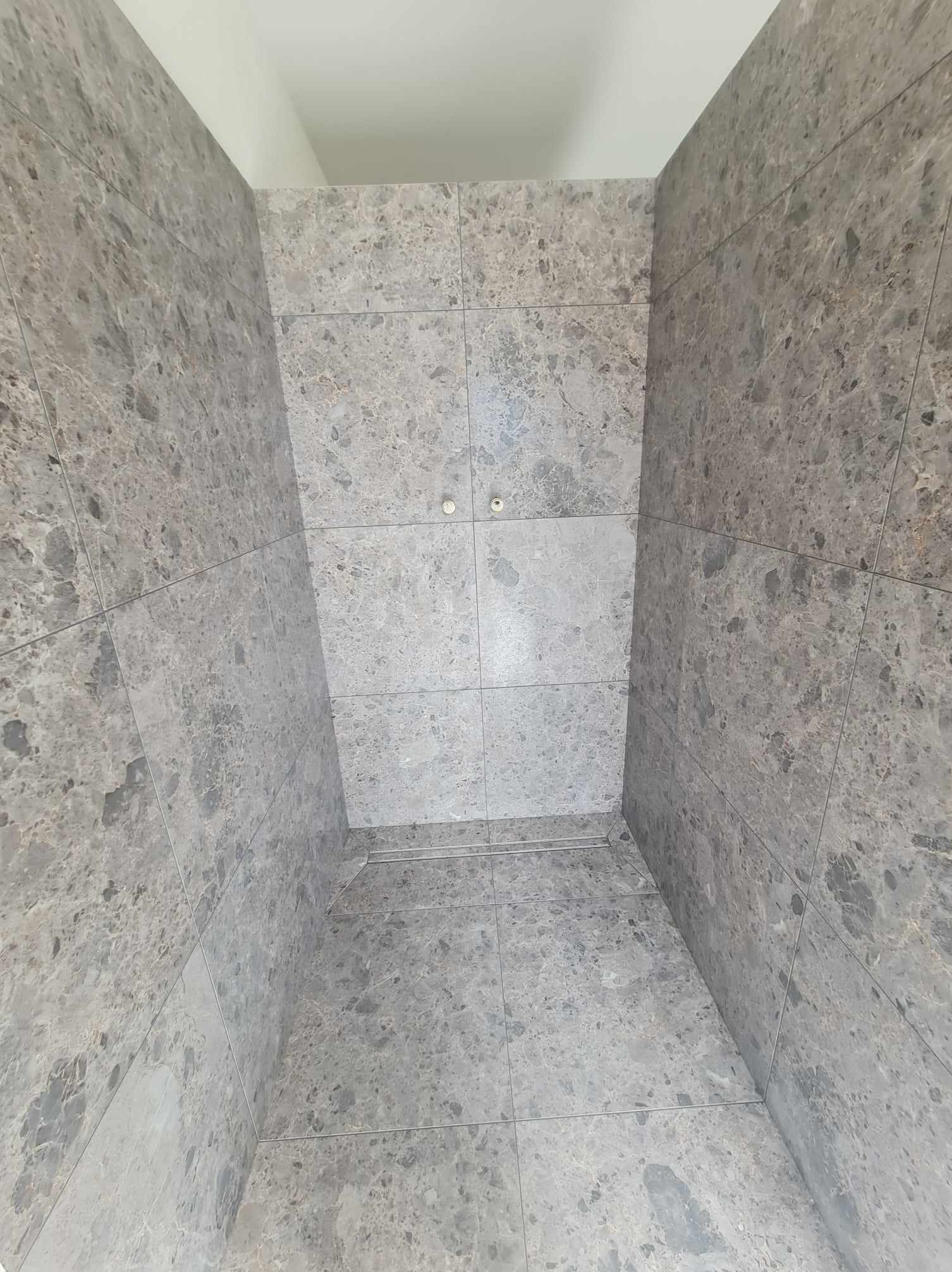 Wykończona kabina prysznicowa z szarymi, marmurkowymi płytkami na ścianach i podłodze, widoczne dwa punkty oświetleniowe w suficie.