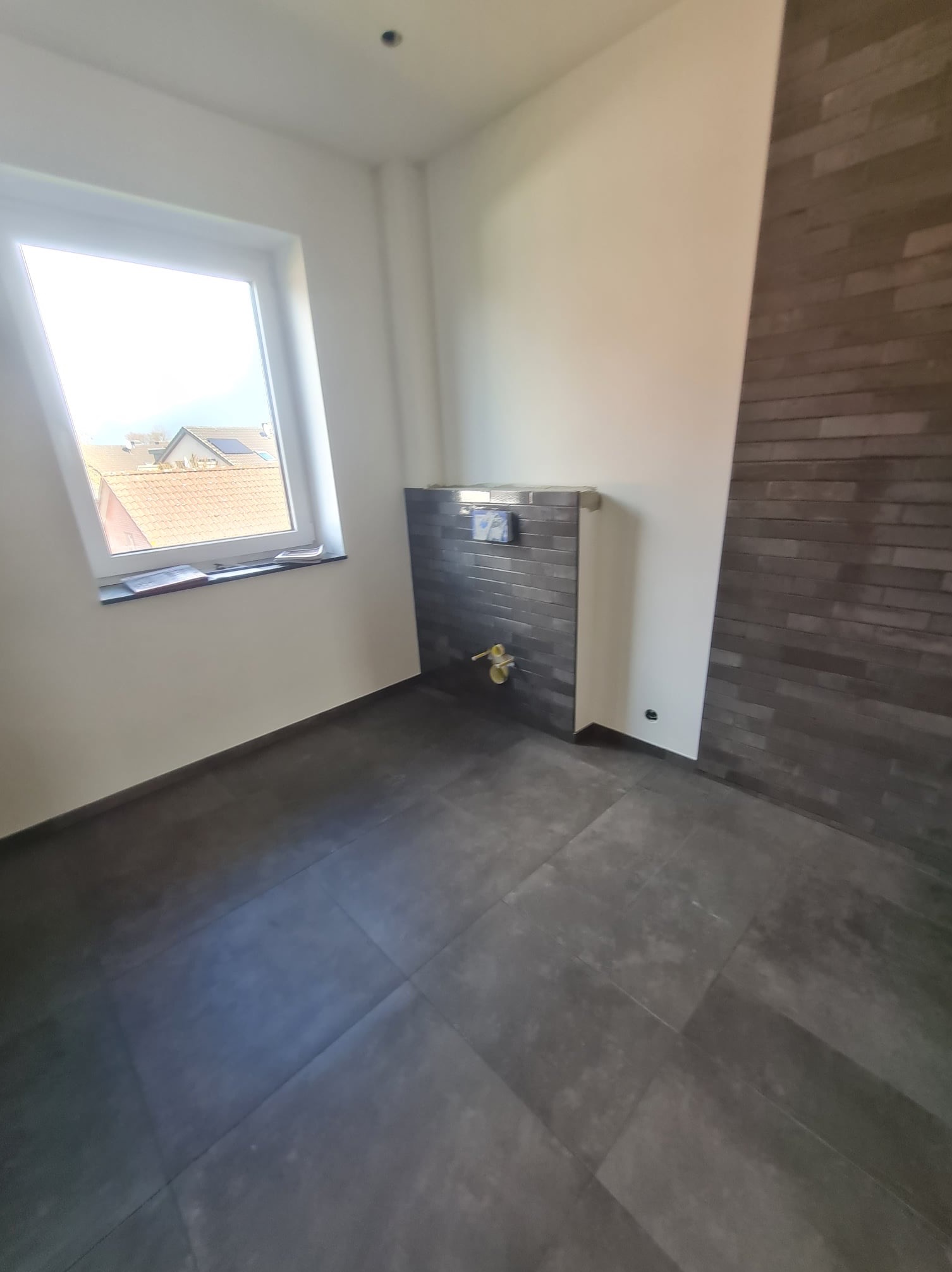 Nowa łazienka w trakcie wykańczania: ciemna podłoga z płytek, fragment ściany z szarych cegiełek, zabudowa podtynkowego stelaża WC częściowo obłożona płytkami, widoczne elementy instalacji wodnej...