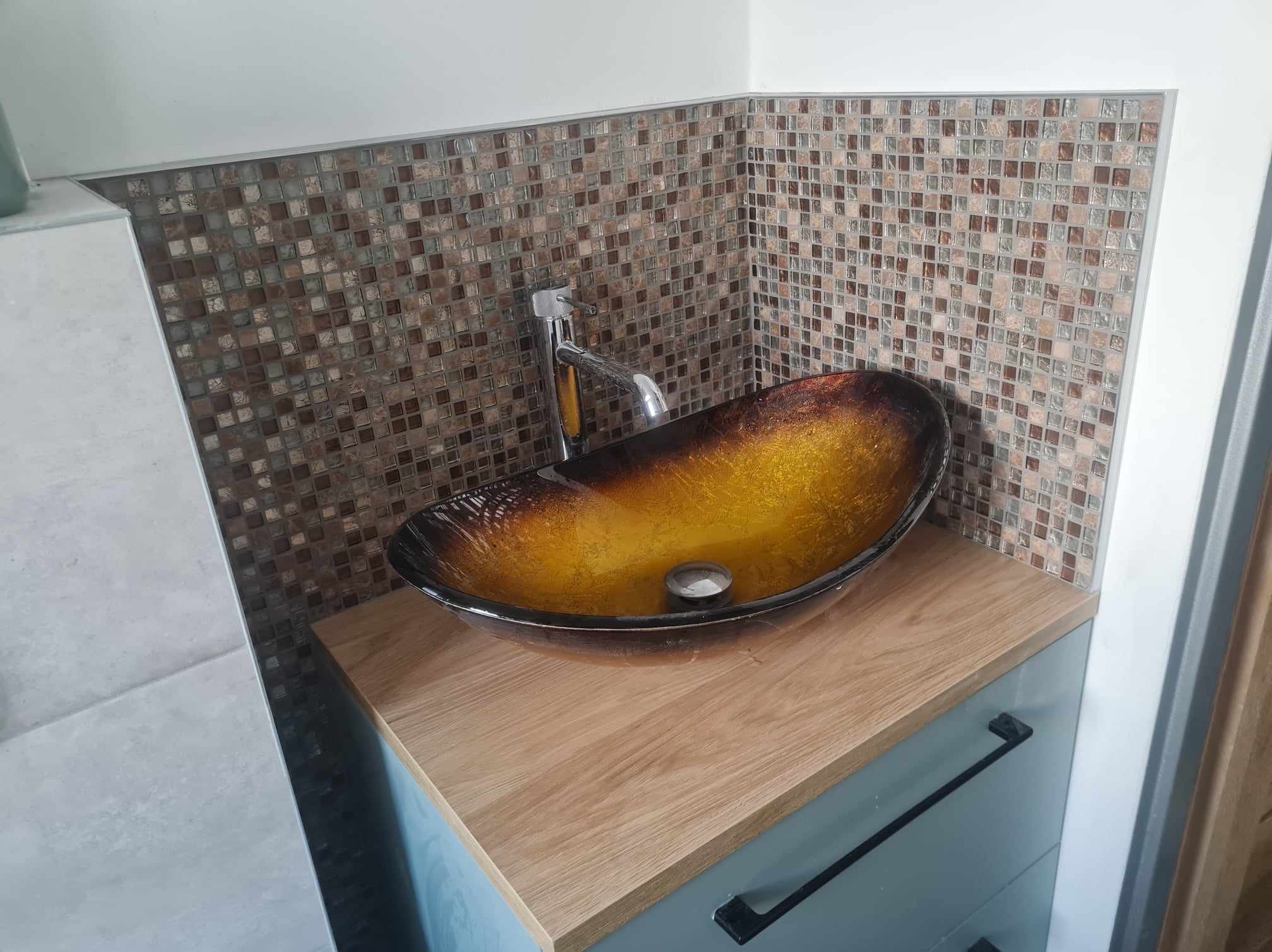 Umywalka szklana w odcieniach brązu i złota, zamontowana na szafce z blatem imitującym drewno, z mozaiką w tle, oświetlona jasnym światłem.
