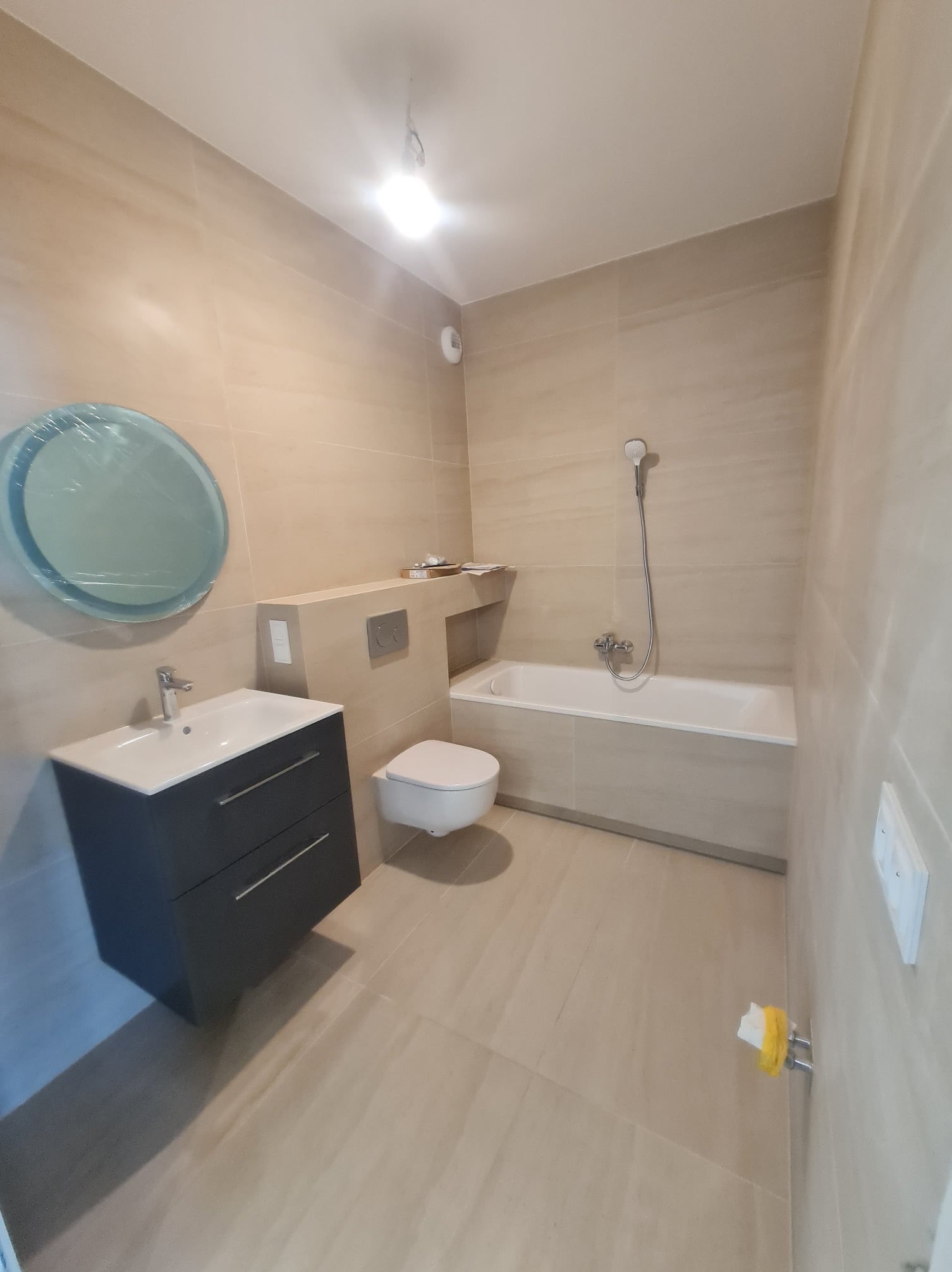 Nowoczesna łazienka z beżowymi płytkami, wiszącą toaletą, umywalką z szafką w kolorze grafitowym i okrągłym lustrem w folii ochronnej.