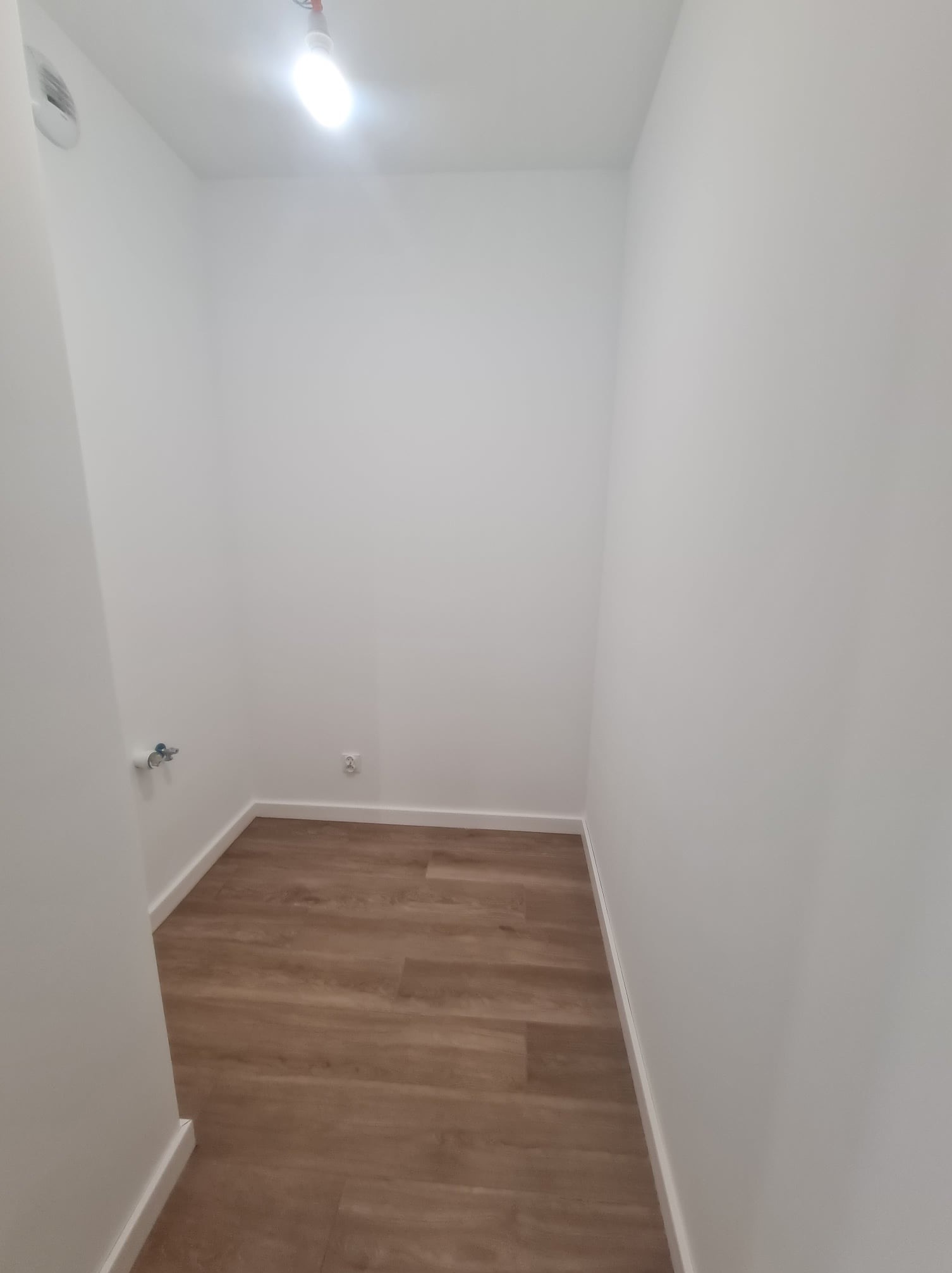 Wnętrze małego, pomalowanego na biało pomieszczenia z drewnianą podłogą; widoczny włącznik światła i gniazdko elektryczne.