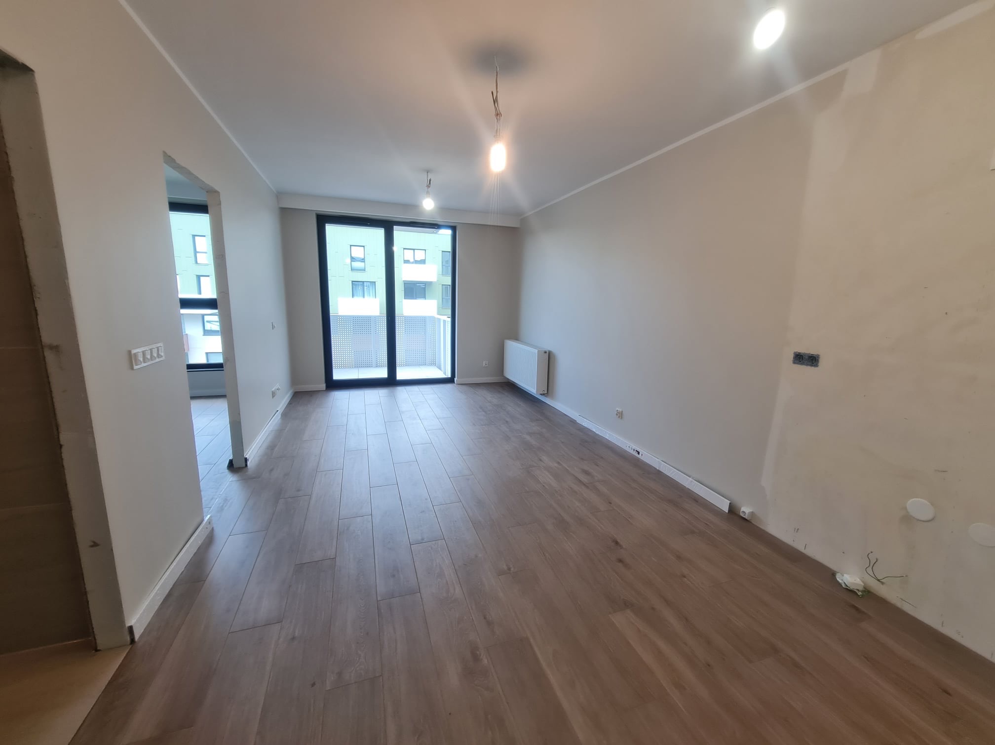 Wnętrze pokoju w stanie deweloperskim z drewnopodobną podłogą, surowymi ścianami z widocznymi śladami prac, grzejnikiem pod oknem balkonowym i otwartym przejściem do kolejnego pomieszczenia.