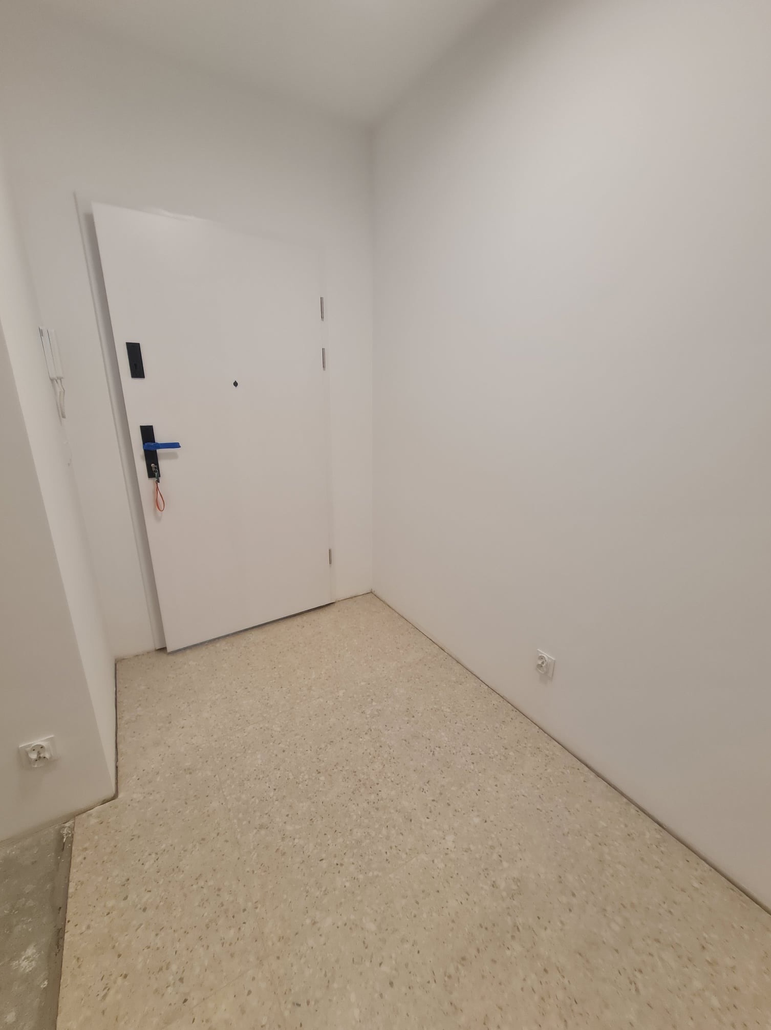 Pomalowane na biało wnętrze przedpokoju z beżową podłogą lastryko i białymi drzwiami wejściowymi z czarną klamką i niebieskim akcentem.