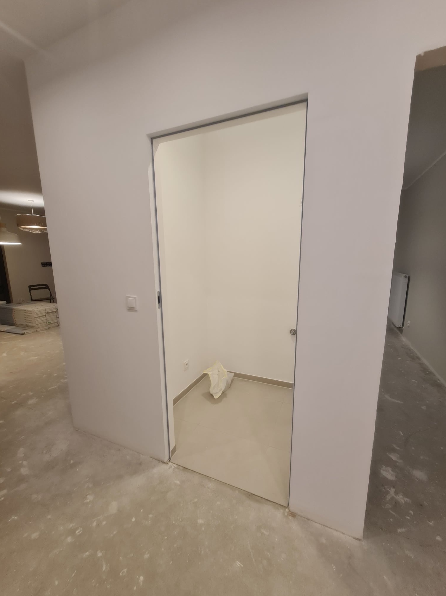 Wnętrze po remoncie z widocznymi świeżo wygładzonymi ścianami w odcieniach bieli, otwarte drzwi do małego pomieszczenia z workiem na podłodze, widoczny fragment podłogi przed i za drzwiami.