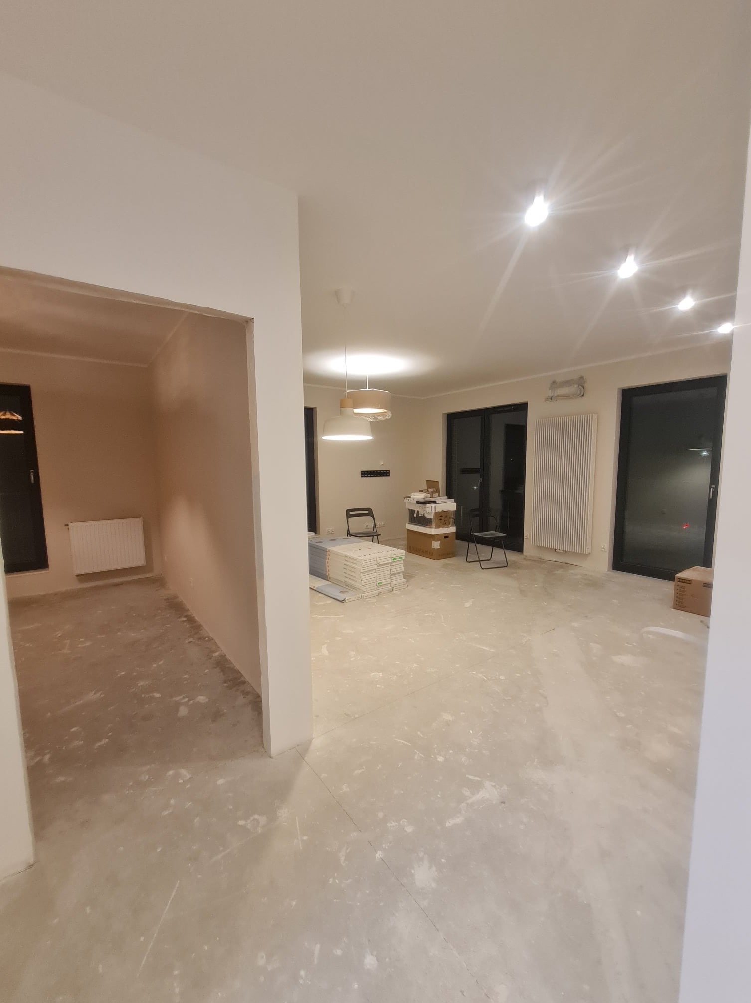 Wnętrze w trakcie prac wykończeniowych, widoczne przygotowane ściany, surowa podłoga, grzejniki i okna balkonowe, w tle kartony i płyty gipsowe.