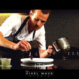 Pixel Wave Studio - Kucharz w trakcie dekorowania talerza sosem z metalowej łyżeczki, obok widoczne pojemniki z ziołami, w tle czarna ściana z mozaiki.