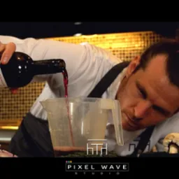 Kamerzysta filmuje ujęcie z nalewaniem czerwonego wina z butelki do miarki, na tle kuchennego blatu z grzybem i logo Pixel Wave Studio w dolnej części kadru.