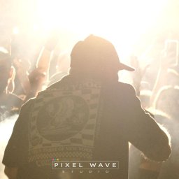 Pixel Wave Studio - Osoba w czapce tyłem do kamery na koncercie, tłum w tle, jasne światła i dym sceniczny, logo na kurtce.