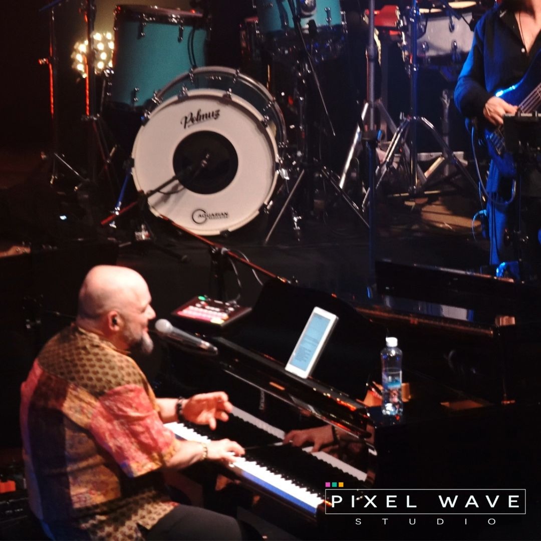 Muzyk grający na fortepianie na scenie podczas koncertu, w tle perkusja i gitarzysta basowy, butelka wody na fortepianie i tablet z nutami.