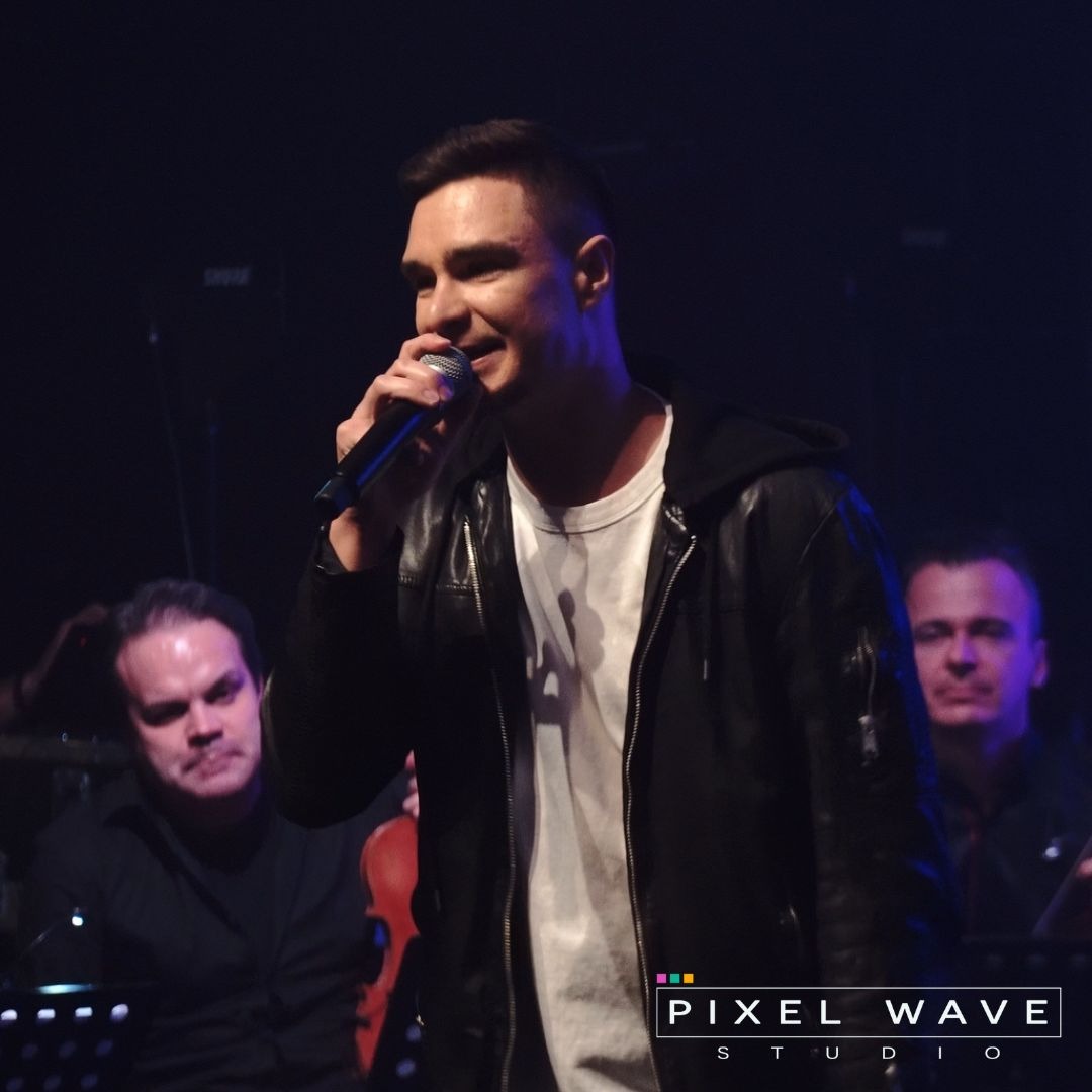 Młody mężczyzna w skórzanej kurtce śpiewa do mikrofonu na scenie, w tle orkiestra smyczkowa i logo studia.