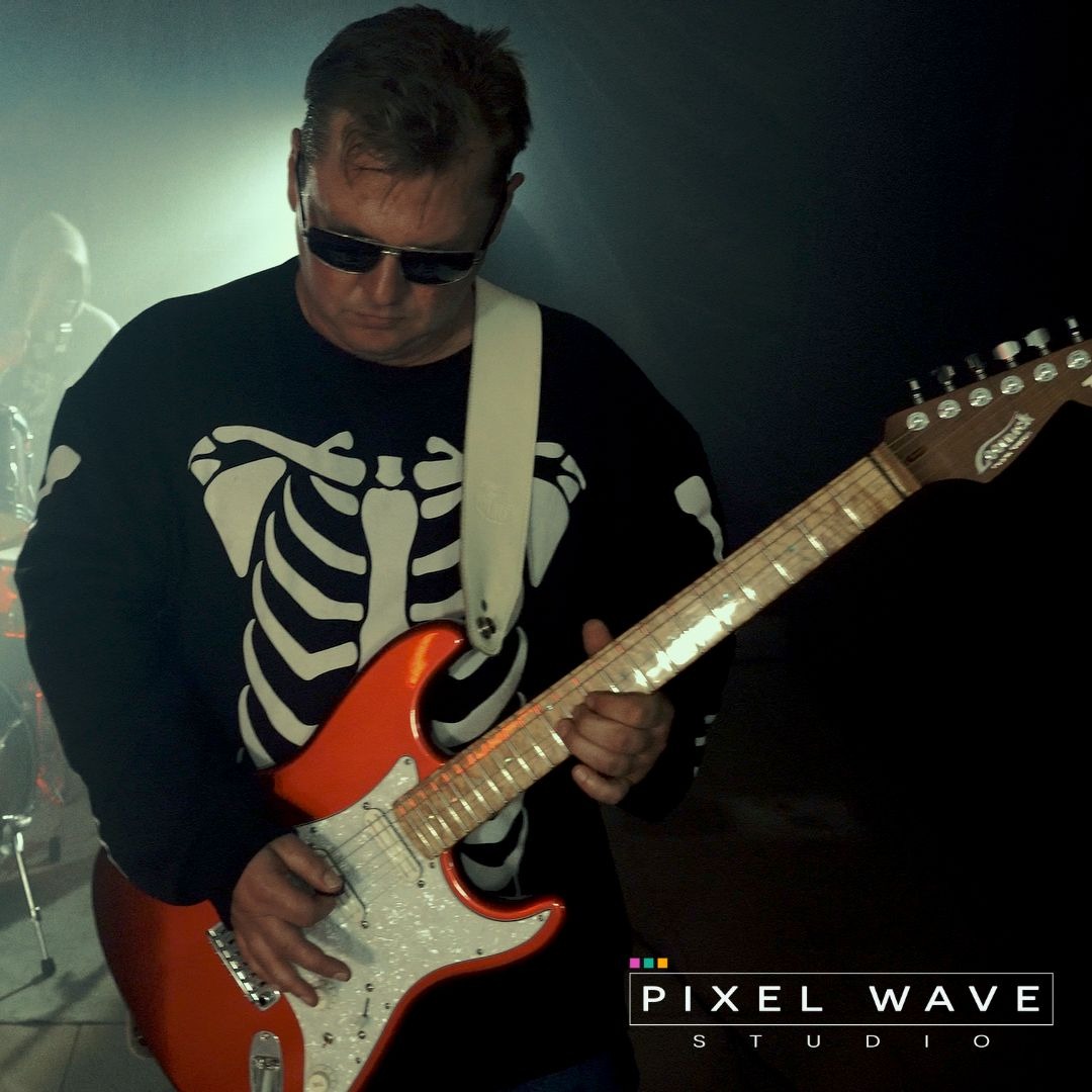 Mężczyzna w okularach przeciwsłonecznych i bluzie ze szkieletem gra na pomarańczowej gitarze elektrycznej w zadymionym pomieszczeniu, w tle perkusja i logo Pixel Wave Studio.