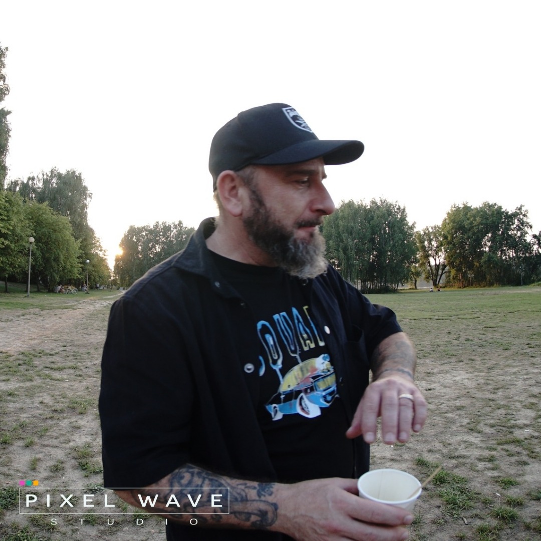 Mężczyzna z brodą i tatuażami, w czapce i koszulce z nadrukiem, trzymający kubek z napojem na tle parku o zachodzie słońca.