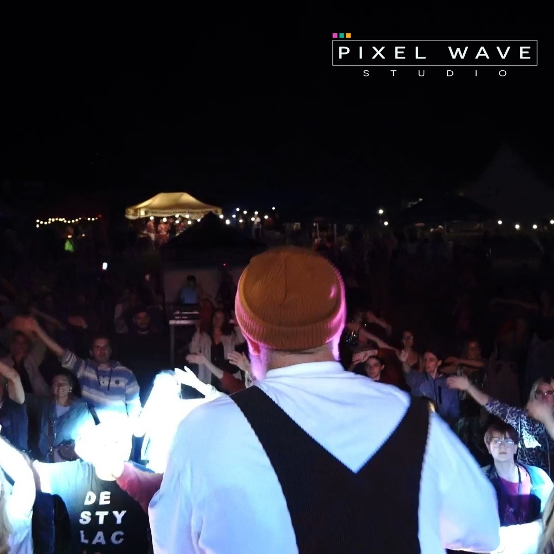 Ujęcie zza pleców osoby w brązowej czapce i białej koszuli z czarnymi szelkami, stojącej na scenie podczas koncertu plenerowego w nocy, z widocznym tłumem i oświetloną sceną w tle.