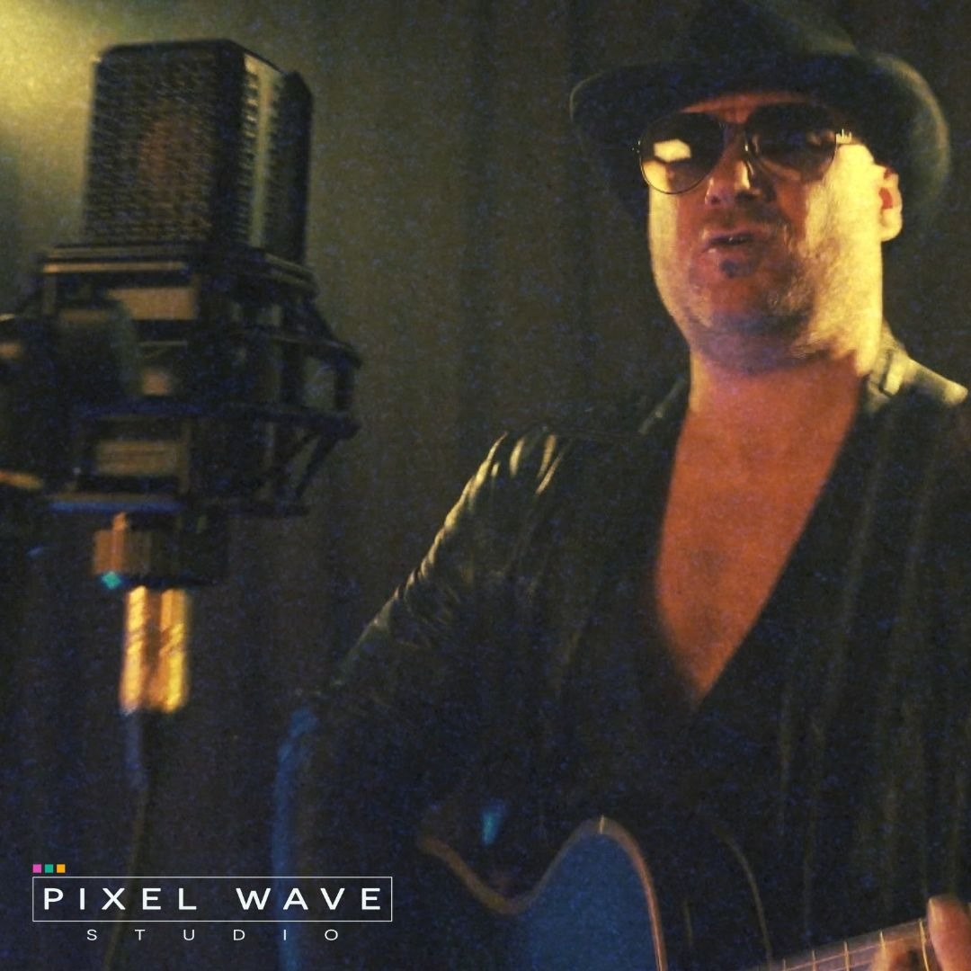 Mężczyzna w kapeluszu i okularach śpiewa i gra na gitarze przy profesjonalnym mikrofonie studyjnym, w tle ciemne tło, widoczne logo studia 'Pixel Wave'.