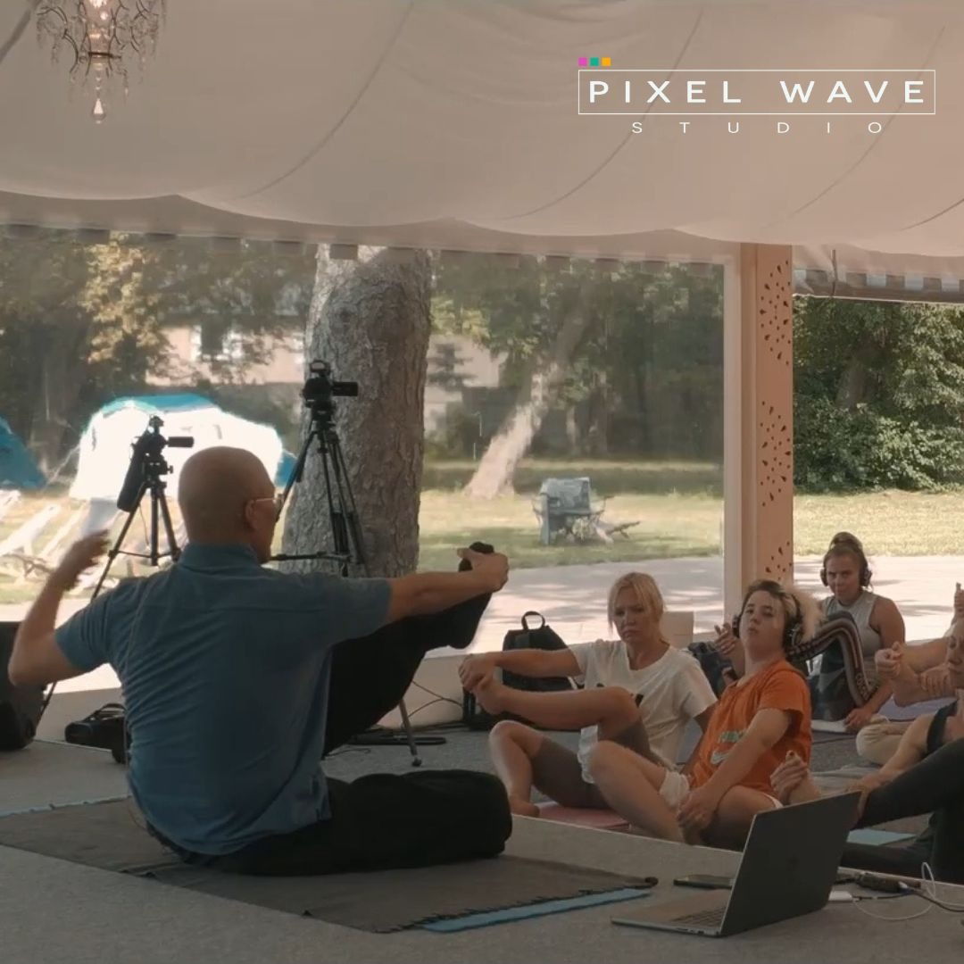 Szkolenie z jogi filmowane kamerą na statywie, uczestnicy siedzą na matach w plenerze pod zadaszeniem, widoczny laptop i logo studia filmowego.