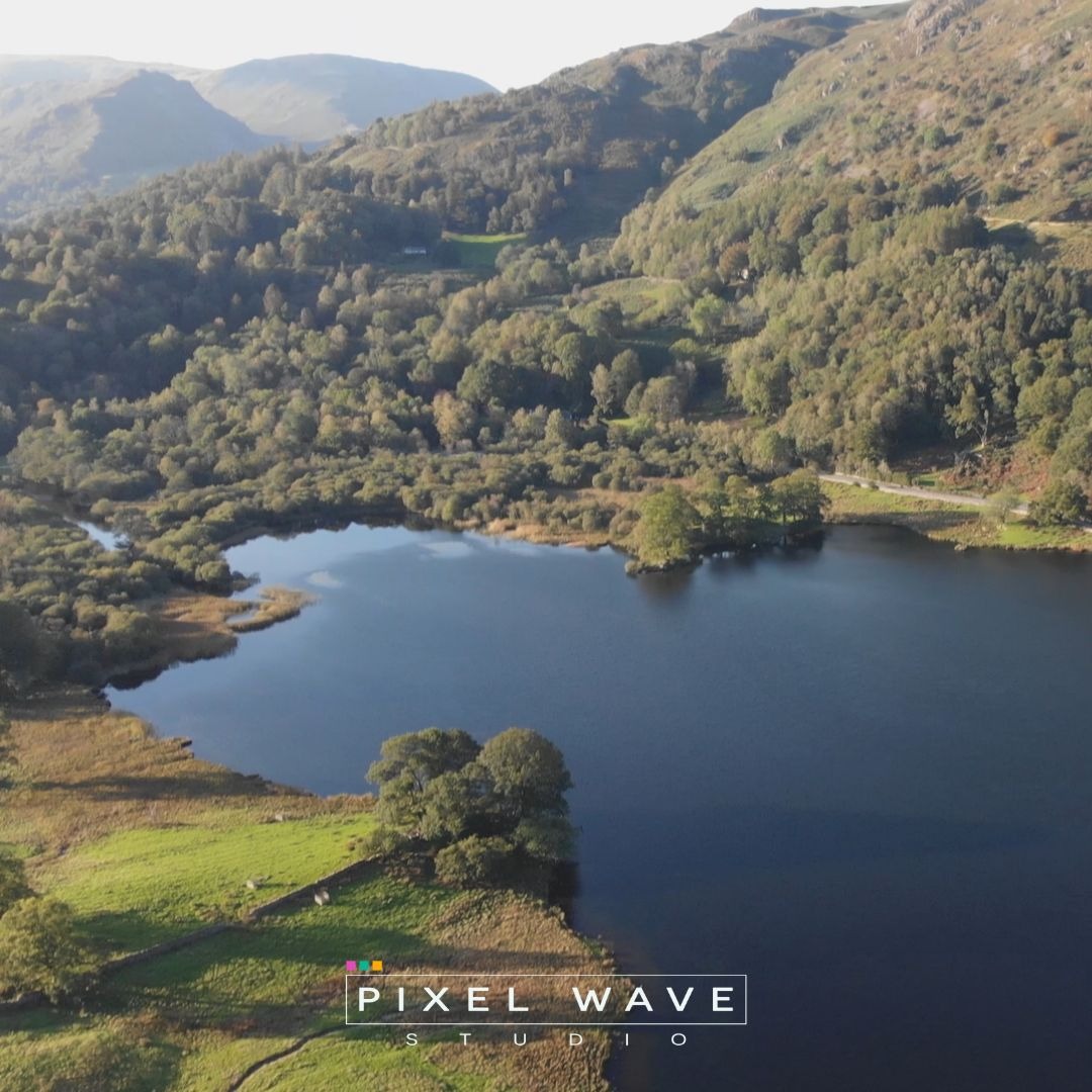 Panoramiczny widok z lotu ptaka na jezioro otoczone zielonymi wzgórzami i lasami, sfotografowane w Komornikach, z logo studia w dolnej części kadru.