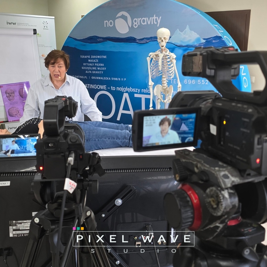 Ujęcie z planu filmowego: dwa profesjonalne kamery w zbliżeniu, rejestrujące sesję terapeutyczną z udziałem kobiety i pacjenta na tle baneru 'no gravity' i modelu szkieletu.