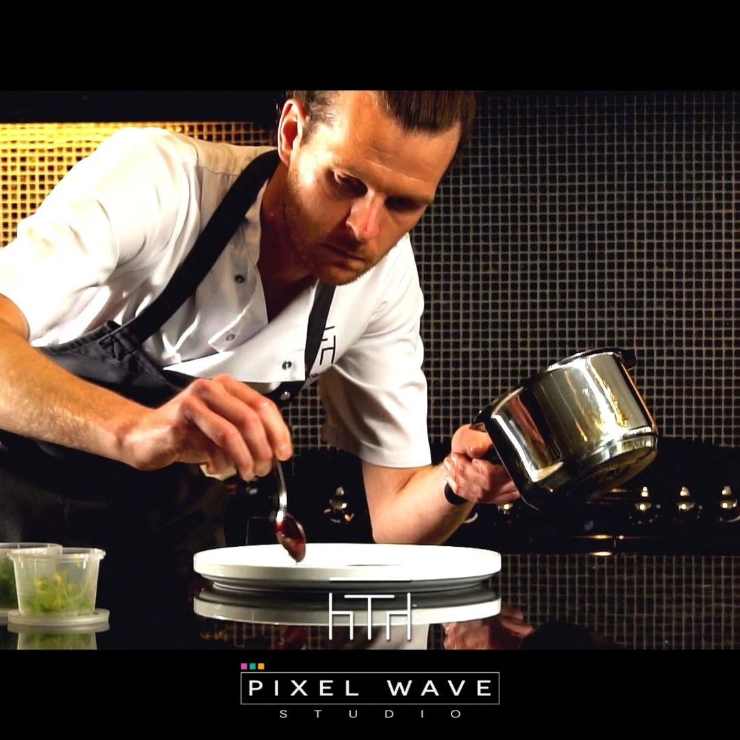 Kucharz w trakcie dekorowania talerza sosem z metalowej łyżeczki, obok widoczne pojemniki z ziołami, w tle czarna ściana z mozaiki.