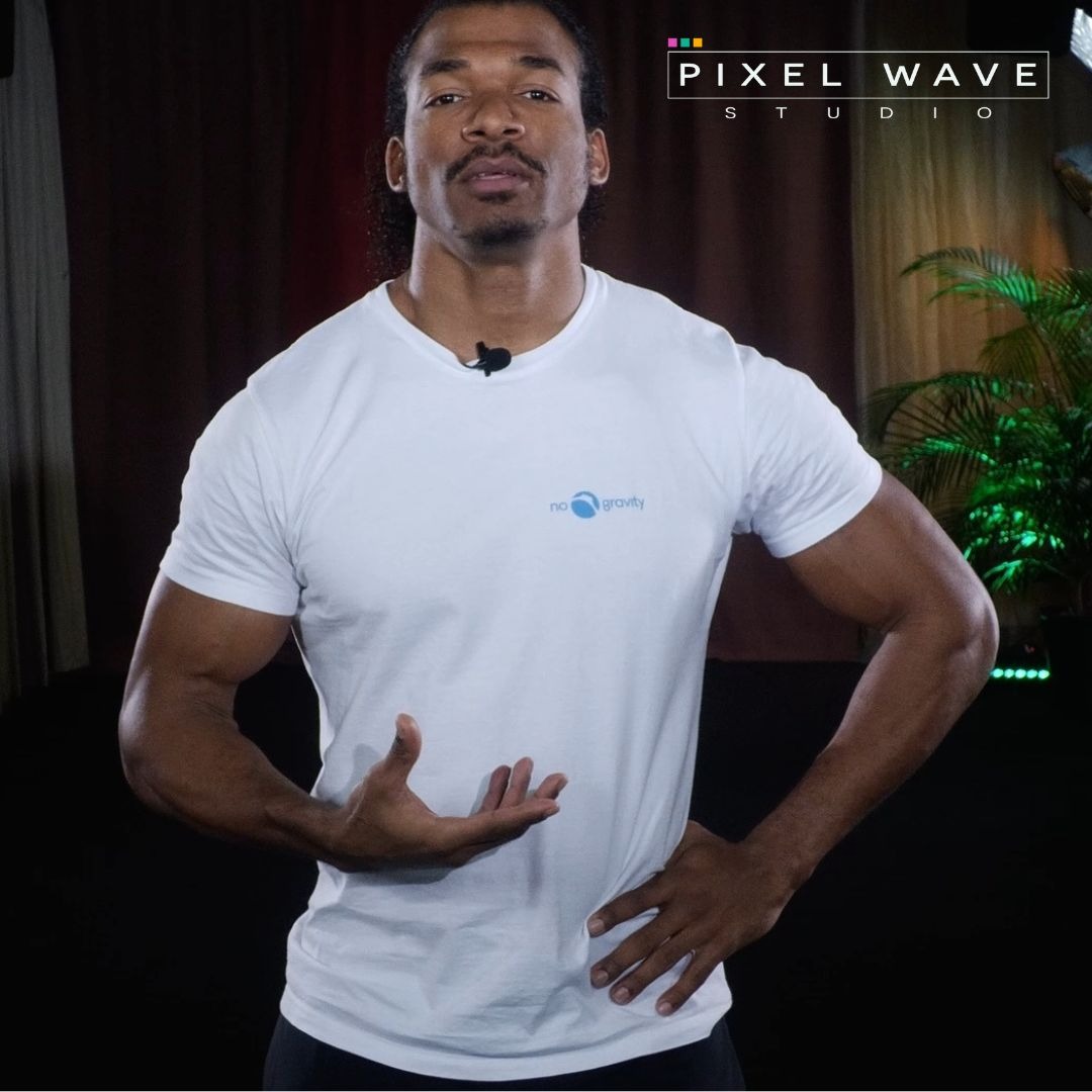 Mężczyzna w białej koszulce z logo 'no gravity' prezentujący gest dłoni na tle studia z rośliną i logo 'Pixel Wave Studio'.