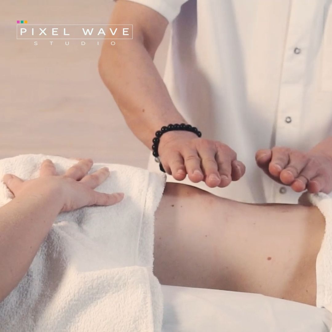 Ujęcie z góry: terapeuta w białym uniformie z czarną bransoletką unosi dłonie nad brzuchem pacjenta leżącego na białym ręczniku.