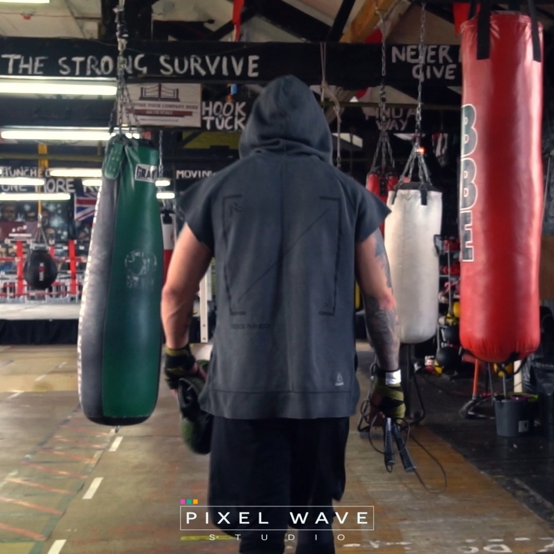 Mężczyzna tyłem w bluzie z kapturem, w rękawicach bokserskich, trzymający skakankę w sali treningowej z workami bokserskimi w tle, pod napisem 'The strong survive'.