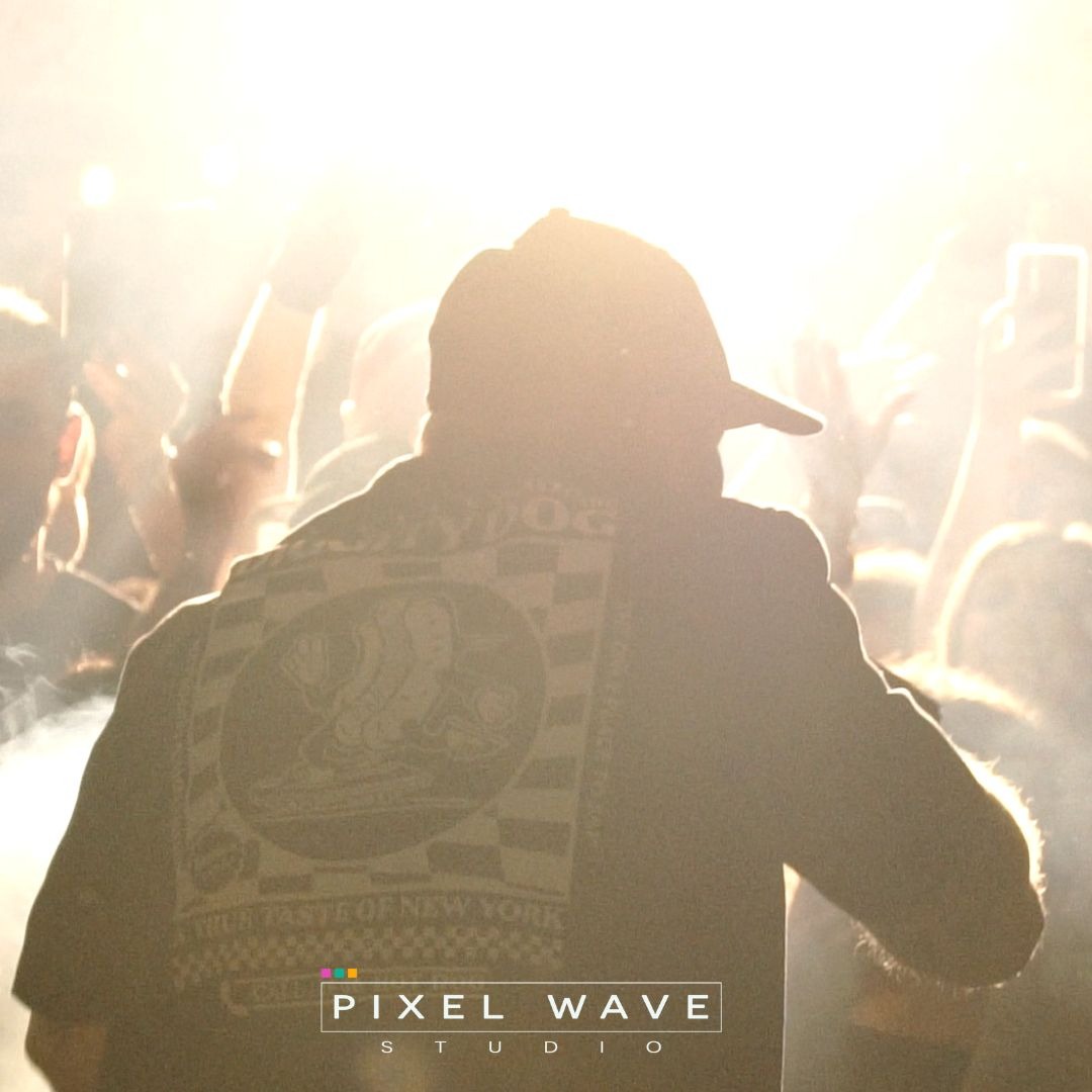 Osoba w czapce tyłem do kamery na koncercie, tłum w tle, jasne światła i dym sceniczny, logo na kurtce.