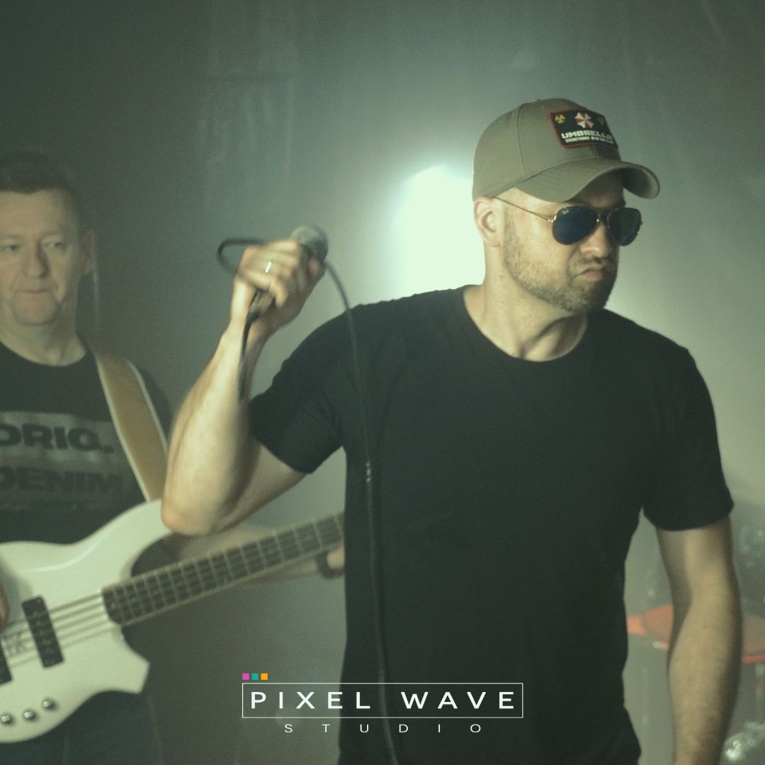 Zespół muzyczny podczas występu na scenie, wokalista w czarnej koszulce i okularach przeciwsłonecznych śpiewa do mikrofonu, basista w tle gra na białej gitarze basowej.