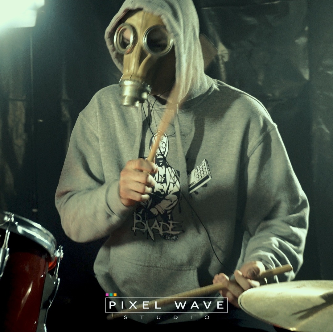 Osoba w masce gazowej i szarej bluzie z kapturem gra na perkusji w ciemnym pomieszczeniu, widoczne logo Pixel Wave Studio.