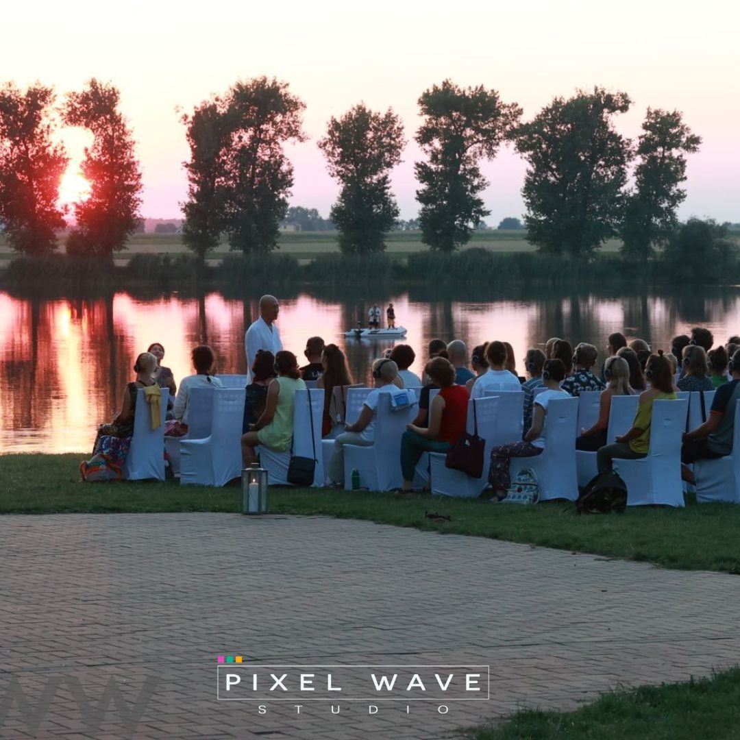 Plenerowa ceremonia ślubna nad jeziorem o zachodzie słońca, goście siedzą na białych krzesłach, w tle para młoda na łódce.