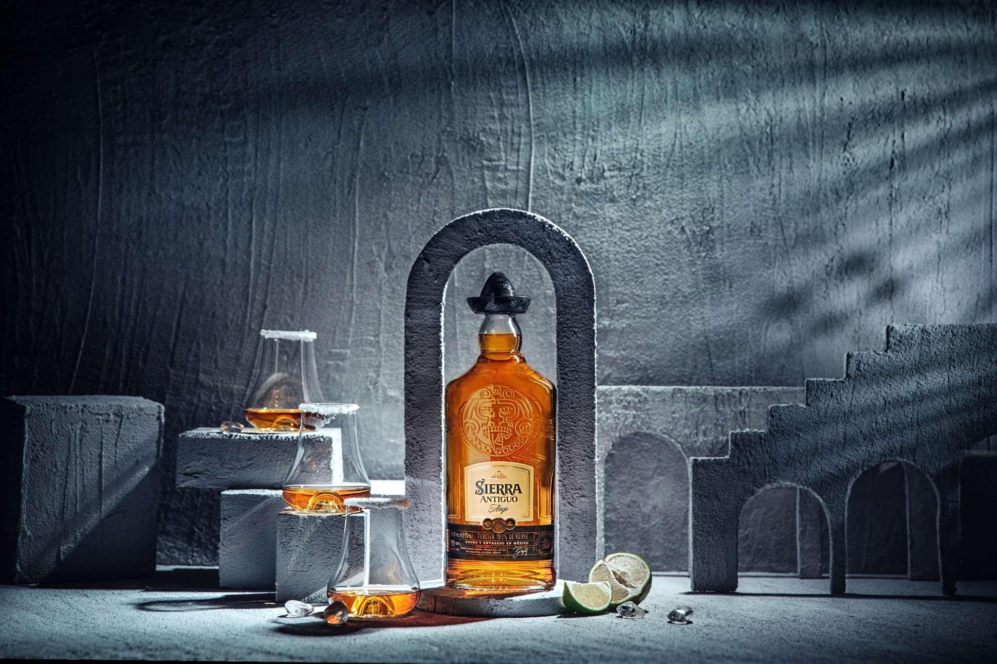 Butelka złocistej tequili Sierra Antiguo w małym sombrero, prezentowana w minimalistycznej scenografii z betonowych bloków, łuków i kieliszków z solą na krawędziach, uzupełniona limonkami.