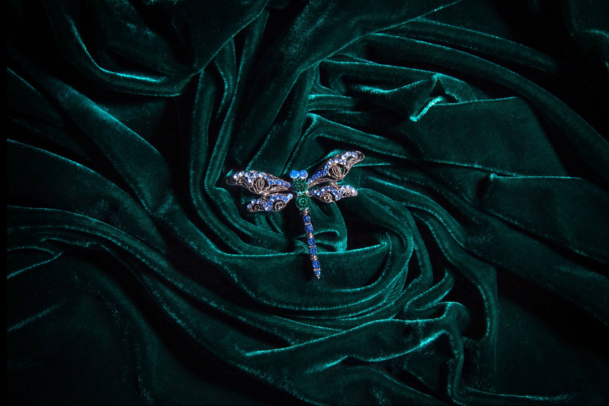 Dekoracyjna broszka w kształcie ważki, wysadzana niebieskimi i zielonymi kamieniami, leży na pofałdowanym, ciemnozielonym aksamicie.