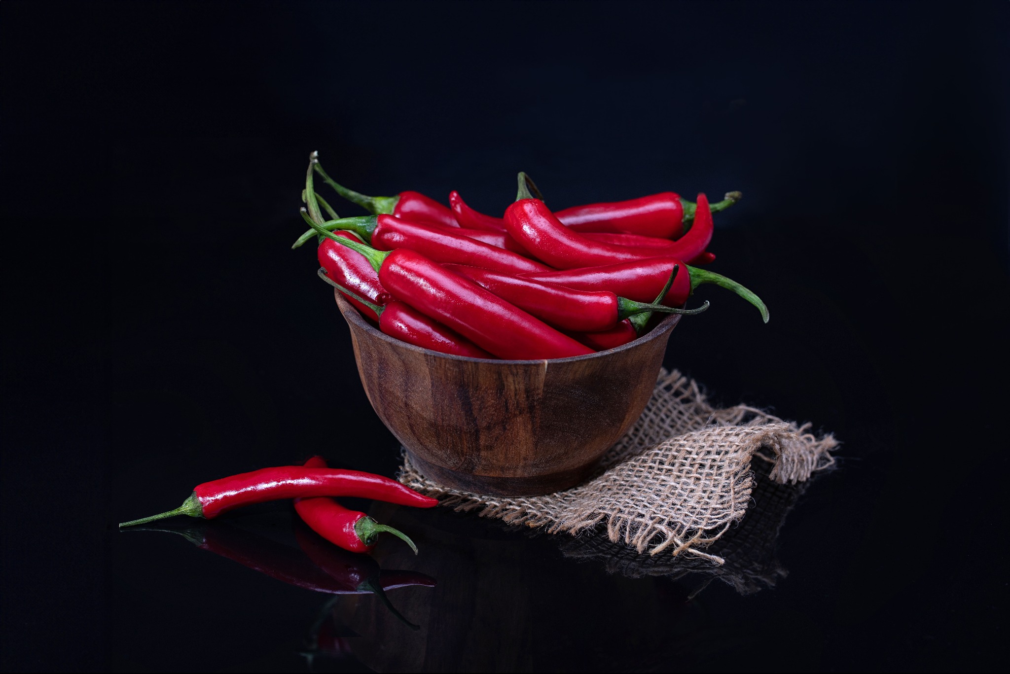 Czerwone papryczki chili w drewnianej misce na jutowym materiale, odbijające się na czarnym, błyszczącym tle.