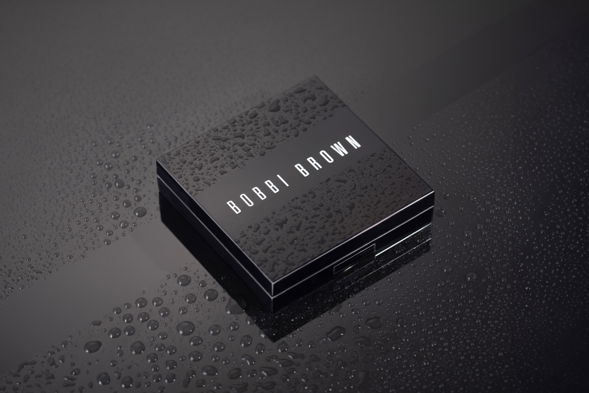 Czarna paleta do makijażu marki Bobbi Brown, pokryta kroplami wody, leży na błyszczącej czarnej powierzchni również pokrytej kroplami wody.