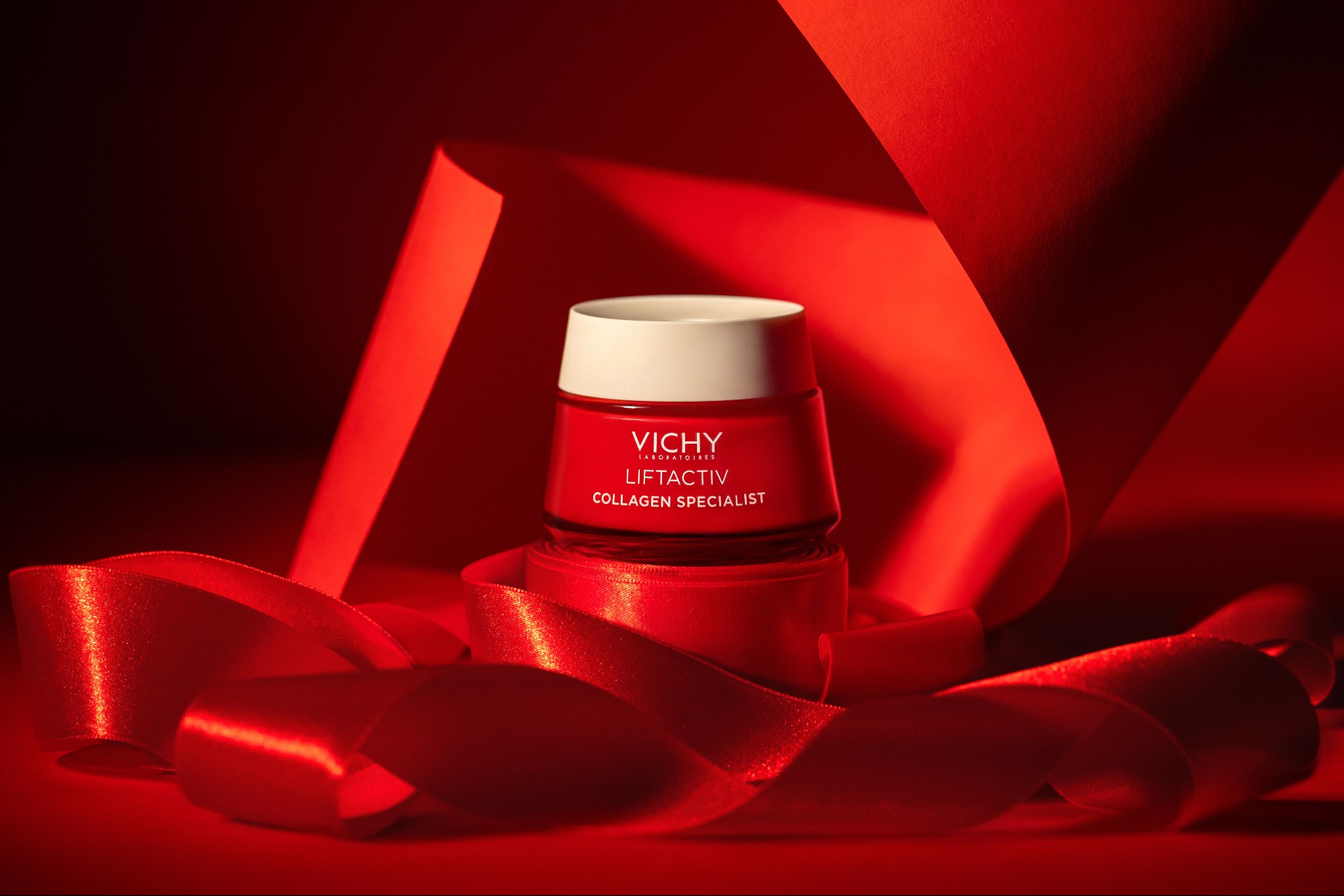Krem Vichy Liftactiv Collagen Specialist w czerwonym opakowaniu, otoczony błyszczącą, czerwoną wstążką na tle z czerwonego papieru.