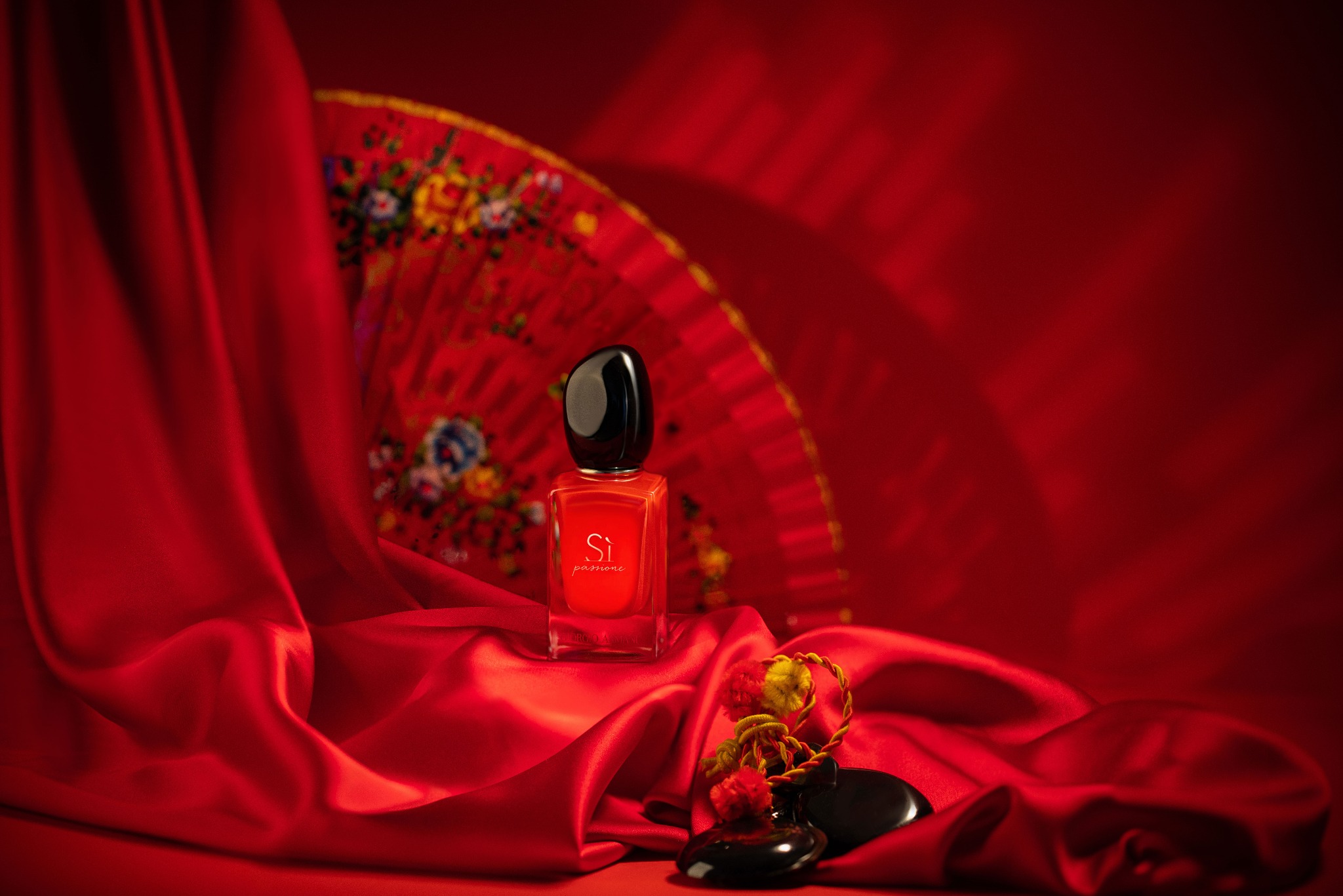 Czerwony flakon perfum Si Passione na jedwabnej tkaninie z wachlarzem w tle, oświetlony studyjnie.
