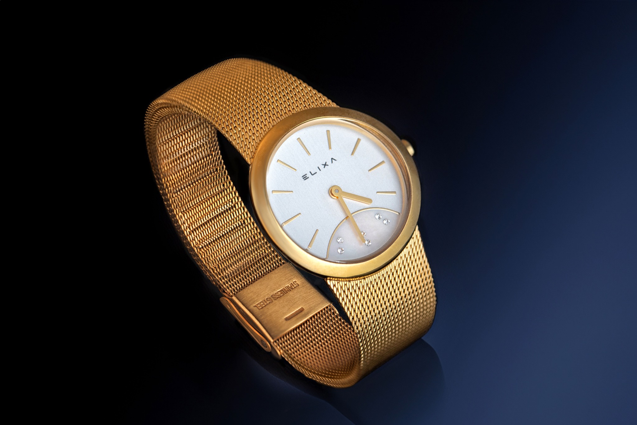 Złoty damski zegarek Elixa z bransoletą typu mesh na ciemnym, niebieskim tle.