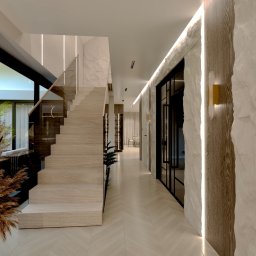 Design Atelier - Nowoczesny hol z drewnianymi schodami i szklaną balustradą. Ściany wykończone kamieniem z oświetleniem LED. Na podłodze jasny parkiet ułożony w jodełkę. Dekoracyjne trawy w wazonie.