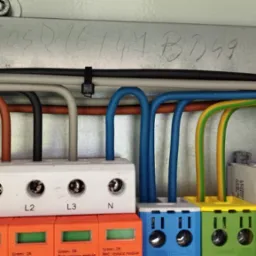 Zbliżenie na podłączone przewody w skrzynce elektrycznej, widoczne kolorowe zabezpieczenia SIMTEC z diodami sygnalizacyjnymi stanu.