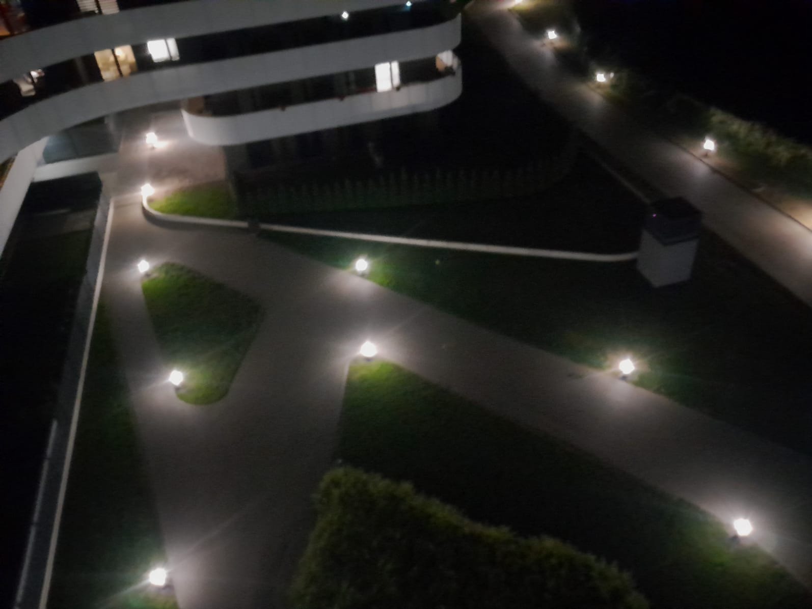 Nocny widok z góry na oświetloną ścieżkę w ogrodzie przy nowoczesnym budynku z balkonami, z wykorzystaniem niskich lamp ogrodowych.