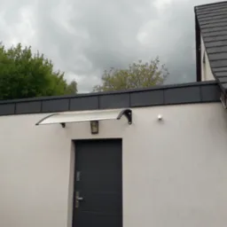 Szara brama garażowa z zamontowanym daszkiem z przezroczystego tworzywa i czarnymi wspornikami, widok na elewację budynku z szarą blacharką dachową i pochmurnym niebem w tle.