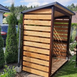 Roof Top - Pogotowie Dekarskie - Drewniana konstrukcja altany z poziomymi listwami, brązowe metalowe słupki, widok z boku na tle zielonego trawnika i ogrodzenia z siatki.