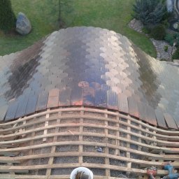 Roof Top - Pogotowie Dekarskie - Układanie dachówki łuskowej na drewnianej konstrukcji dachu o nieregularnym kształcie, widok z góry na częściowo pokryty dach, narzędzia pracy w pobliżu.