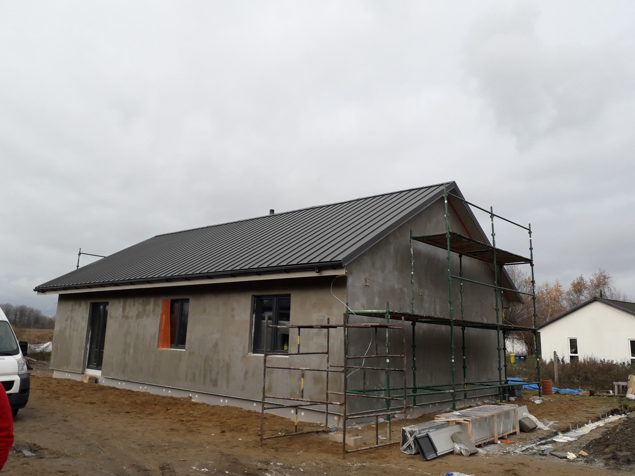 Dom jednorodzinny w trakcie budowy: szare ściany zewnętrzne, ciemny metalowy dach, rusztowania przy ścianie, widoczna warstwa ocieplenia pod dachem.